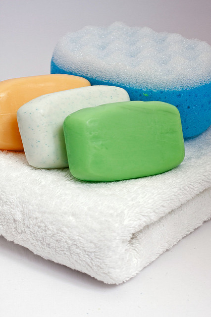 Three bars of soap of orange, white and green colors, next to a double sided bathing sponge with a porous texture. All these hygiene items are on top of a folded white towel made from a fluffy fabric.